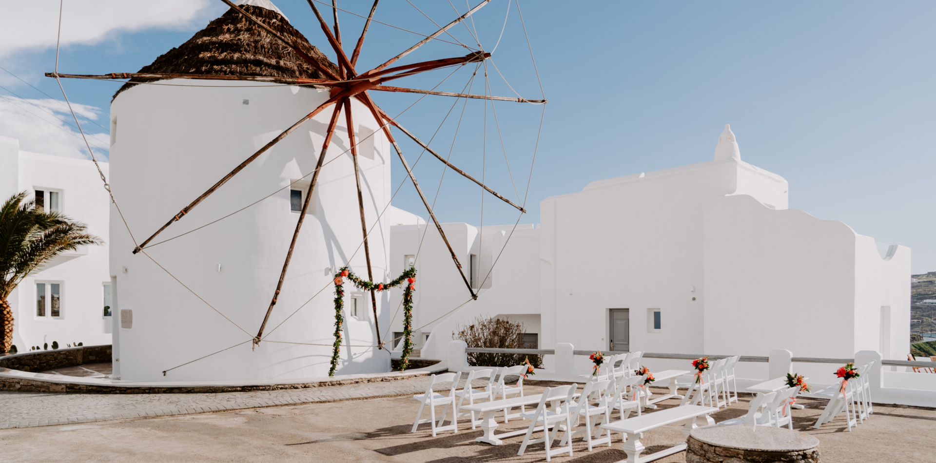 A wedding ceremony setup at the iconic windmills in Mykonos, using orange flowers and greenery as wedding decoration, perfect for a Mykonos wedding.