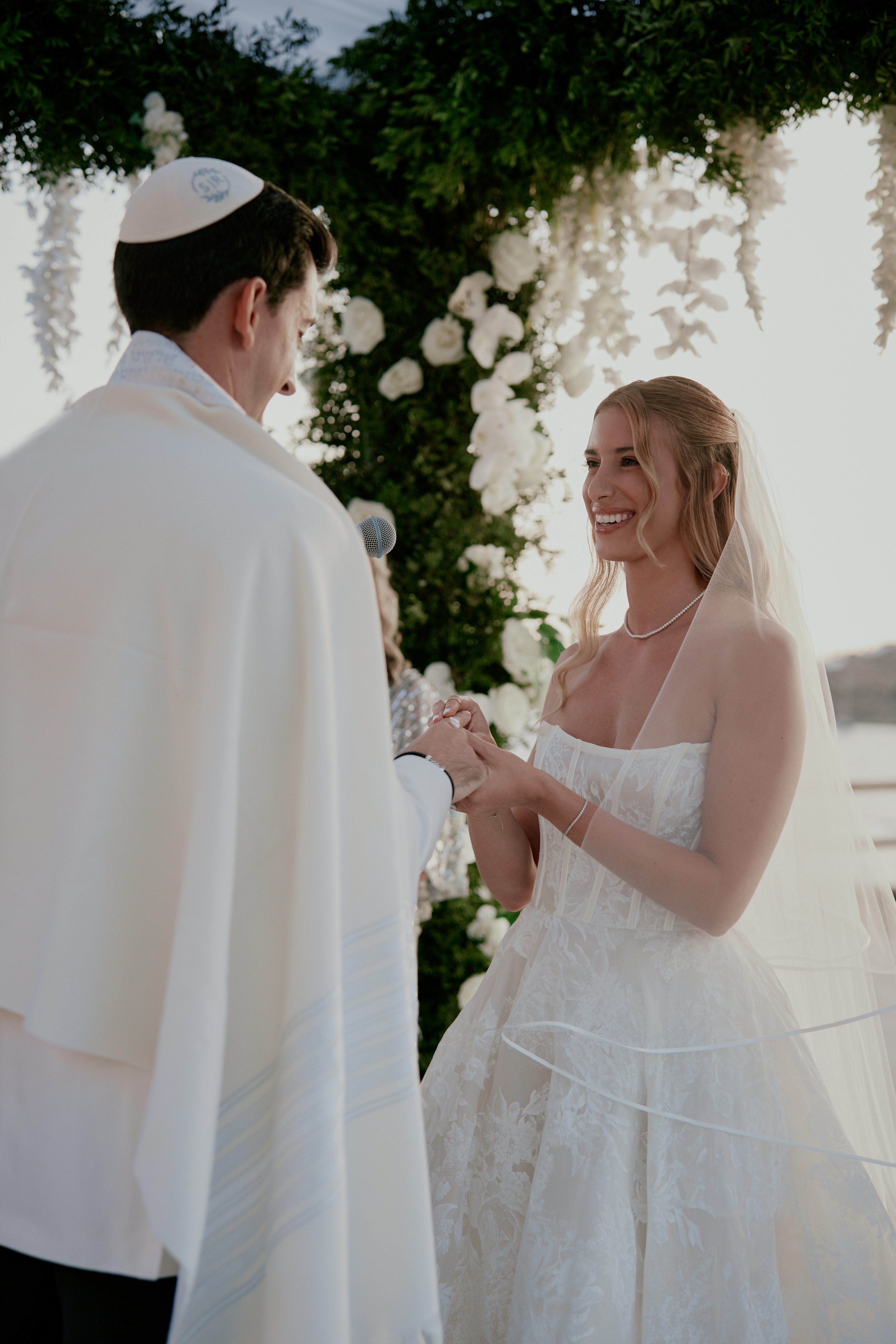 Bride and groom exchanging rings under a lush floral chuppah during a romantic luxury Jewish wedding in Mykonos with the sea glistening behind them