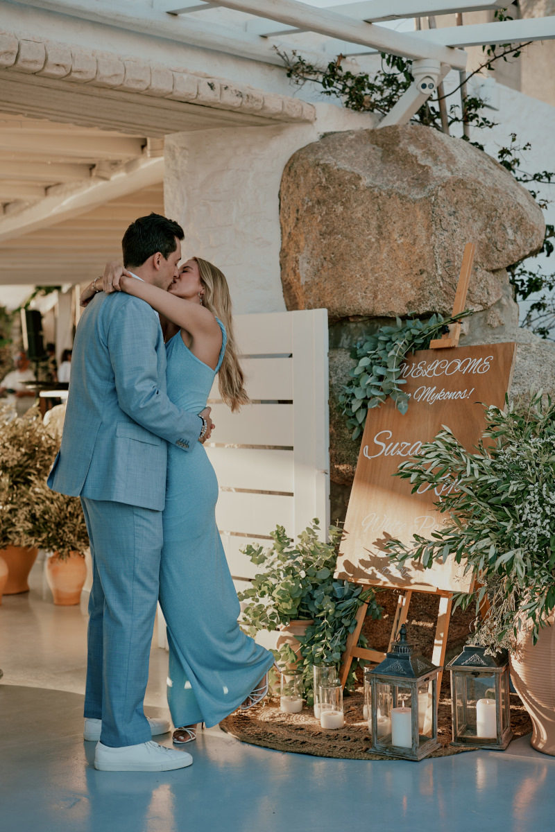 A couple kissing at the entrance of their wedding venue, welcoming their guests for their Greek themed pre wedding white party in Mykonos.