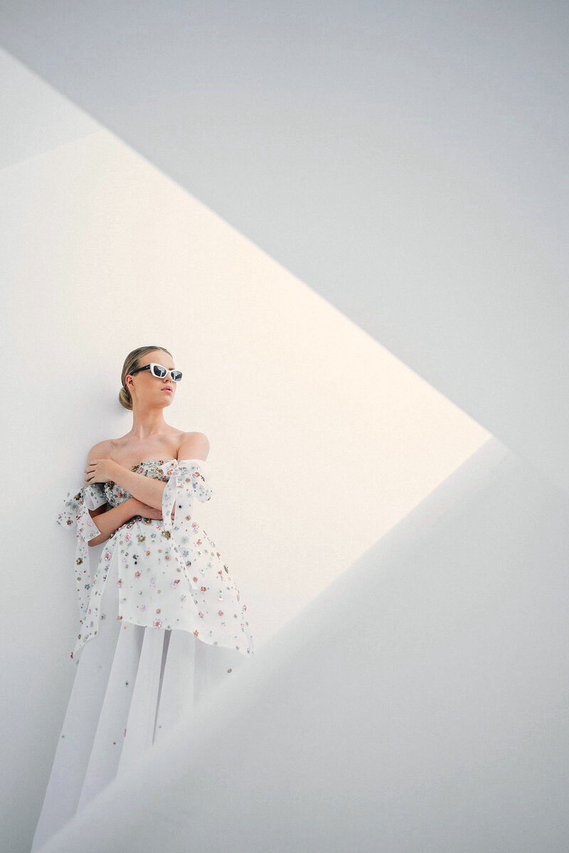 Stylish bride in a floral wedding dress and sunglasses posing in modern white architecture — luxury wedding weekend in Mykonos planned by full-service wedding planners