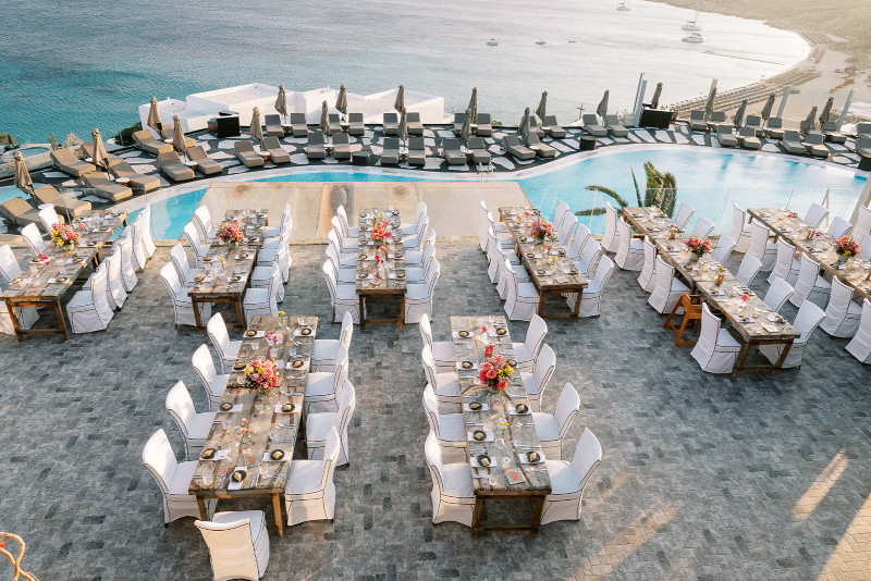 A Luxury Mykonos wedding reception set up by the pool beside dinner tables with flower table centerpieces and chairs.