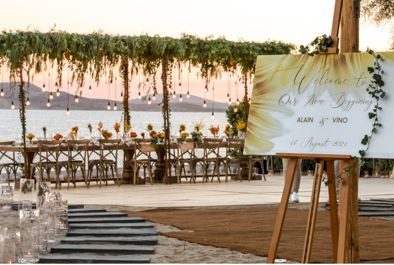 A Mykonos wedding reception set up on a beach, with a custom design structure on top of a dinner table with flower centerpieces